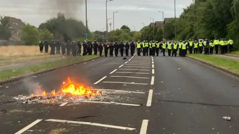 BBC/Alison Blackstone A photo of a mid-size single carriageway road. About 20 metres away is a line of police in neon yellow or black uniforms, walking away from the photographer. About 5 metres away is a pile of debris on fire in the road.