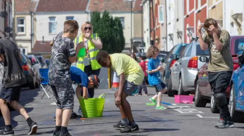 A group of children have a water fight on a suburban street on a sunny day. A woman in a yellow high-vis jacket is supervising. Cars line the street.