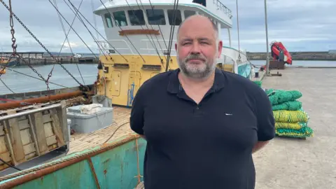 A man stands in front of a large beam trawler on the main quay in Newlyn harbour