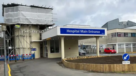 Outside the main entrance of Princess Elizabeth Hospital in Guernsey. A flower bed with no plants in it is outside the main entrance. Parts of the building has scaffolding.