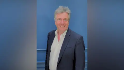 Association of Dental Groups Neil Carmichael has grey hair and is wearing a white shirt, open at the neck, and a navy blazer. He is standing against a blue background.