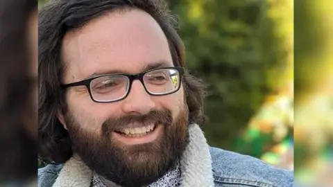 A bearded man wearing glasses. He is wearing a fleeced denim jacket.