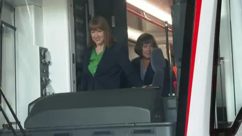 Rachel Reeves with Jo Stevens in the cab of a tram train, being seen through a window.
