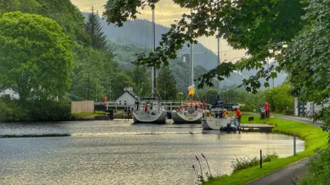 Aileen Gillies A peaceful canal scene featuring several moored sailboats near a lock, framed by lush green trees and rolling hills under a cloudy, golden-lit sky.