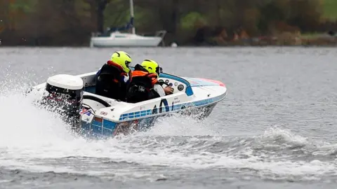 John Heald Photography A blue and white power boat on the water moving at speed. There are two people wearing fluorescent helmets in the boat. There is a white boat in the background.