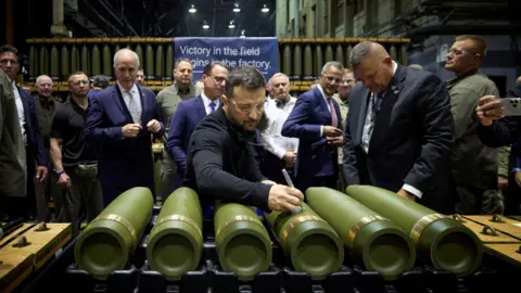 Flanked by other officials, Ukrainian President Volodymyr Zelensky writes with a pen on an artillery shell in a factory