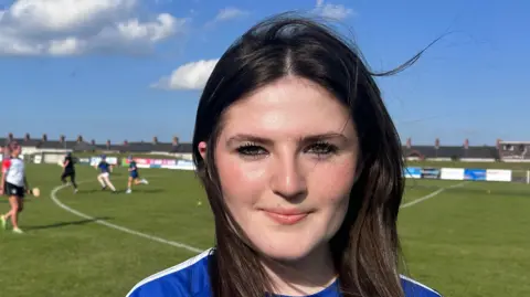 Emma Duffy is standing on a grass pitch as women play football behind her. She has long brown hair and is wearing a dark blue and white trimmed t-shirt.