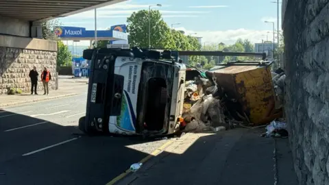 A white lorry cab on its side, with the front window having a circular cut. The skip that it was carrying is on the side with piles of rubbish spilling out. 