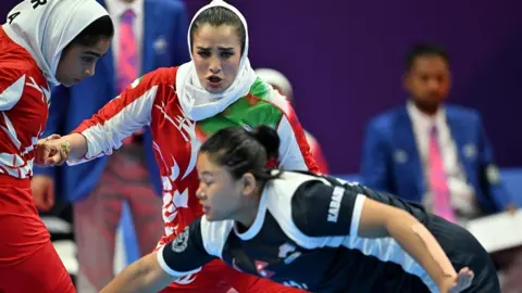 Getty Images Women kabaddi players dressed in red, white and black. The nearest one, with black hair tied back in a bun, is leaning forward while behind her, a woman in red and white with a scarf, tries to reach for her.