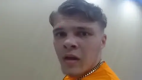 Devon and Cornwall Police Sonny Boyes looking into the body camera with his mouth open. He is wearing a gold chain and yellow top.