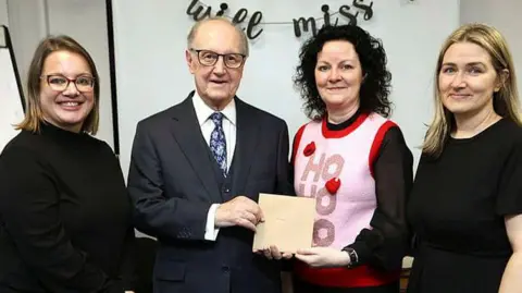 Four people stand together looking at the camera and smiling, three are women, but second left is Mr Harris. He wears a navy blue three piece suit and white shirt, with blue patterned tue. He is holding a card with a brunette lady, who is wearing a pink and red Christmas jumper that reads 'Ho Ho Ho'. The women either side of them are blonde and wear black clothes.