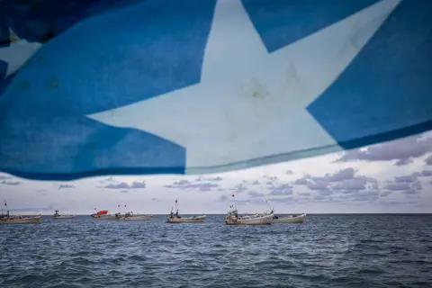 ED RAM / GETTY IMAGES Somalia's national flag - bright blue with a white, five-pointed star in the middle - is blown by seawinds. The background boats are visible.