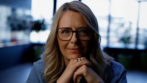 EA A woman with sleek blonde hair and black glasses poses for a portrait in an office setting. She's smiling with her chin resting on her hands.