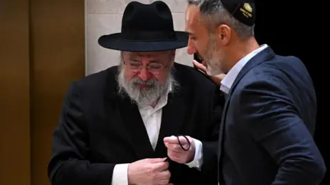 A man with beard and glasses and dressed in a black suit and hat cries and is comforted by a second man wearing a dark suit and kippah