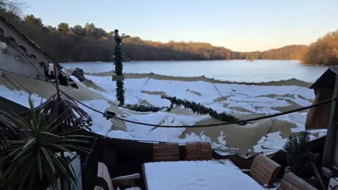 The beige tent has fallen and looks flat next to the building next to it. There are poles with Christmas decorations around them that have pierced through the tent with lots of white snow resting ontop.