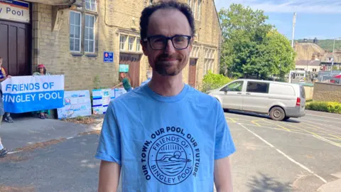 A man in a blue T-shirt which reads 'our town, our pool, our future' and 'friends of Bingley pool'. He is wearing glasses. He is standing in front of a building and there are a few people holding a banner which says 'friends of Bingley Pool'.