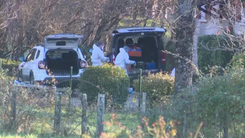 France Television Criminalist agents taking boxes at or from the boots of a car, with another car with its open boot next to them. Trees and bushes are all around them.