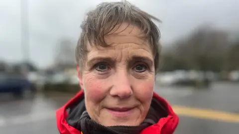 Alwynne Shannon is close to the camera. She's wearing a red coat and has short hair. The background is blurred.