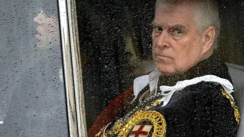 Reuters Prince Andrew sitting behind a glass window in a car with raindrops on it. He is looking glum and wearing a royal uniform with golden trim and a St George's cross on it.