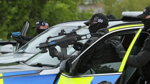 Getty Images Two masked police officers stood beside a white police car, holding guns 