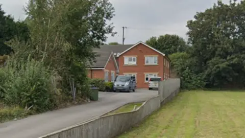 A house at the end of a long driveway. There are trees on one side and a green on the other.