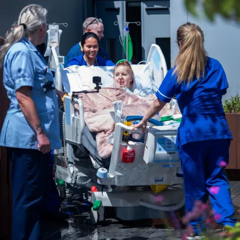 BBC Hollie, 29, a patient at King's College Hospital, is wheeled onto the new rooftop intensive care unit in south London in her hospital bed, surrounded by three nurses dressed in blue uniforms.