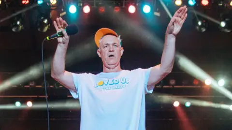 Getty Images A man with grey hair in a pale blue t-shirt and orange cap with a microphone in one hand and his hands in the air, with various stage lights behind him.