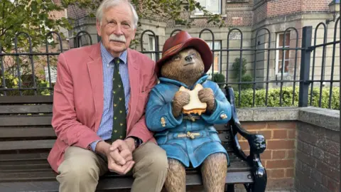 A statue of Paddington sitting on a bench holding a marmalade sandwich with an elderly gentleman smiling beside him