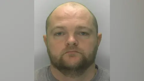 A mug shot of Anthony Williams. He is a balding man with a trimmed beard, wearing a grey jumper
