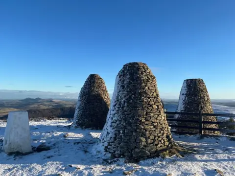 Duncan Hotchkiss The Three Brethren cairns, which consist on three tall mounds of stones, outside Selkirk, with Eildon Hill visible in the background. It's a snowy day with a blue sky.