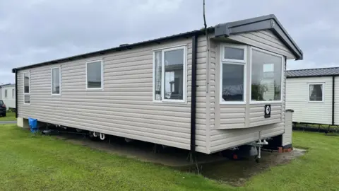 Andrew Dawson The Dawsons' caravan at Skipsea Sands holiday park. It is beige-coloured with white windows and black gutters and a bay window at the front. It is surrounded by grass and other caravans can be seen in the background.