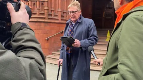 A man stands outside a redbrick building with a large staircase. He has grey hair and glasses. He is wearing a blue overcoat, carrying a walking stick, and reading from a tablet. Two journalists are standing in front of him.