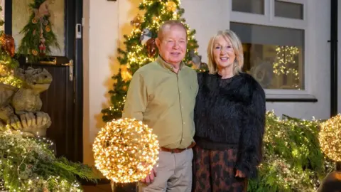 Kirsty Anderson A man wearing a green shirt and woman with blonde hair and black jumper stand in front of a house decorated with Christmas lights and decorations