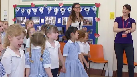 A group of young girls aged between 4 and 7 standing up in their school assembly room watching a former nurse at the front showing them how to rub sun cream in. The girls are dressed in their uniform which is a mix of white t-shirts and skirts and blue and white stipped short-sleeved dresses.
