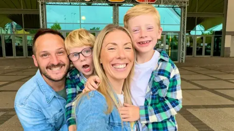 Daisy Woods, pictured second right, smiles in a selfie with her sons Sonny (left) and Oscar (right) and her husband Andy (far left). Both her and Andy are wearing denim shirts, while Sonny and Oscar are wearing matching white t-shirts and green, blue and yellow checked shirts over the the top.