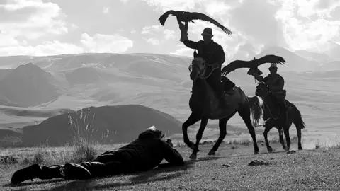 Adam Docker Adam Docker is laying on the ground, he is filming two men on horseback, they are holding up eagles in their hands. The background shows rolling hills and mountains.