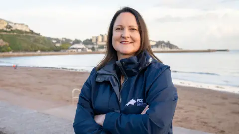 Alison Hernandez stood in front of a beach. She has long brown hair and is wearing a dark blue jacker. 