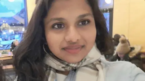 Aarushi A girl with long brown hair is wearing a checkered scarf and puffer jacket. She is standing in a restaurant smiling at the camera.