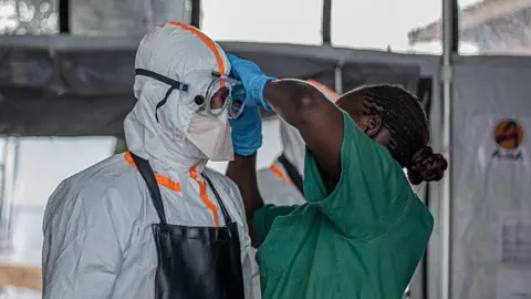 A health worker in green scrubs helps another person put on their PPE. The person on the left of the picture is wearing full white protective gear and goggles.