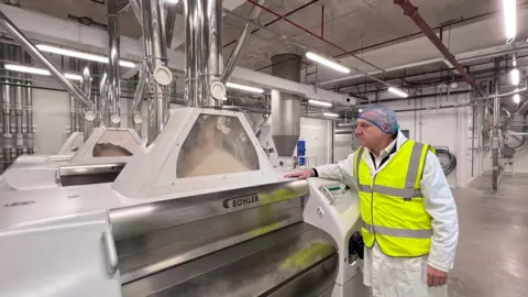 A man watching a big machine in a factory, where flour is behind a clear glass panel on top of it, coming down from a vertical metal pipe. He is wearing a blue hairnet, white lab coat with a yellow high viz over it.