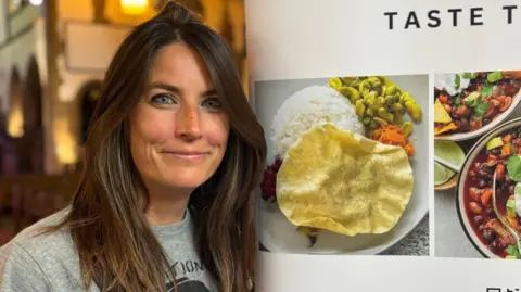 Emma Bamber stands smiling to the camera, next to a banner for her organisation. Pictures of food are seen on the banner. She has long brown hair and blue eyes and is wearing a grey t shirt.