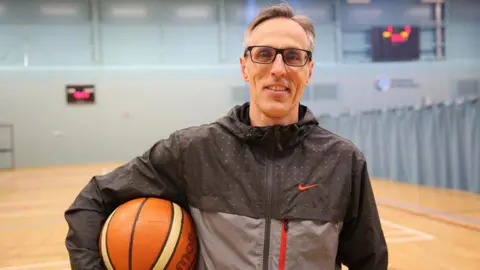 A man in a black and grey Nike zip-up tracksuit top smiles in a sports hall while holding a basketball under his arm. Behind him a sports court can be seen with a curtain and netting dividing the room in half, and digital scoreboards. The man has greying black hair and thick black glasses.