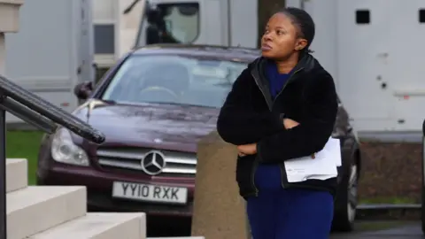 BBC A woman wearing a blue dress and a black jacket has her arms folded as she is walking by the steps to the court.