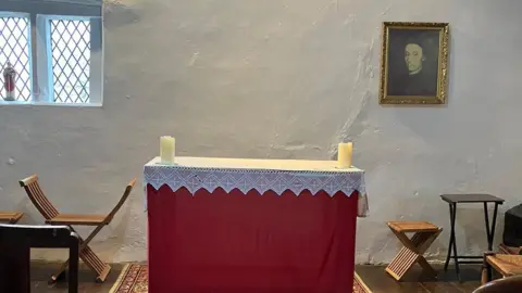 Claire Ashmore/BBC The altar in the attic in Brindle where Edmund Arrowsmith celebrated his last Mass. It has a white lace cover and a red cloth draped down to the floor. There are two candles on it, and a painting of the saint hangs on the wall.