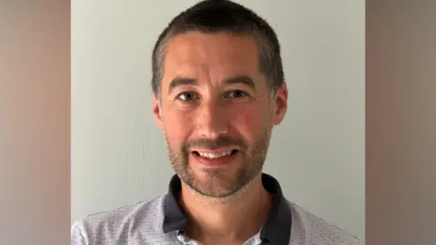 Matthew Bailey A head and shoulders shot of a Councillor Matthew Bailey - a man with short brown hair, black stubble. He is wearing a black and grey polo shirt against a white wall.
