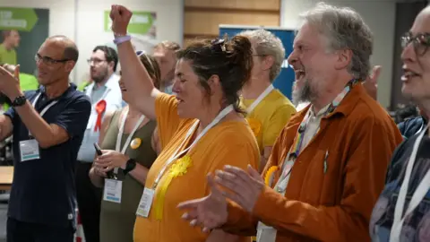 Bethia Thomas, wearing a yellow T-shirt and rosette, celebrates with a fist in the air. A man next to her, also wearing a yellow rosette, is cheering and applauding.
