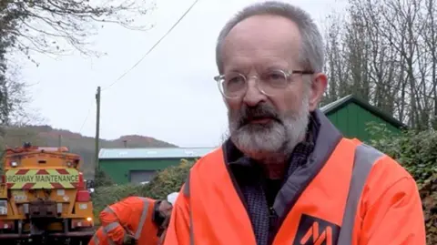 Shropshire Council A man with grey hair, a beard and an orange jacket in front of bushes, a man with an orange jacket and a yellow lorry