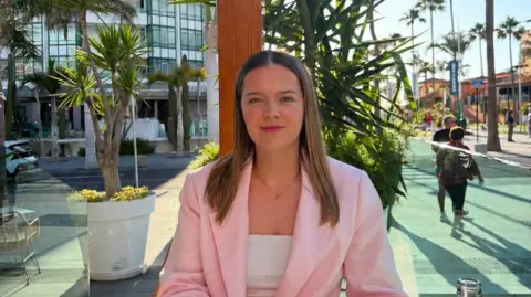 Mia Edgworth Mia is pictured in a sunny climate. She's wearing a light pink blazer with a white top underneath and a dainty necklace. She has light brown hair.