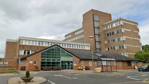 A large brick built hospital with a shelter in front. In front is a large single-storey building with a triangular glass-fronted entrance. Behind it is a three-storey building with long, uninterrupted windows along each floor. To the right is a seven-storey building in the same style, with a large stairwell structure just off-centre. The buildings are arranged in a T-shape with the tallest building at right angles to the other two.