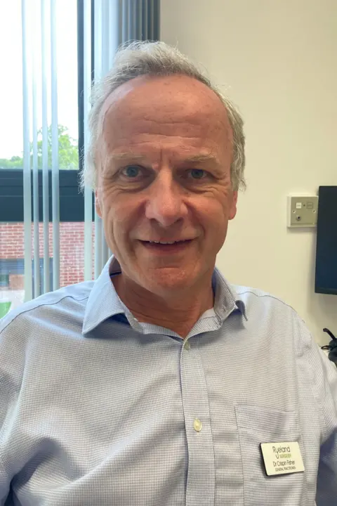 BBC / Alistair Binney A man with grey hair, wearing a white and light blue patterned shirt, wearing a name tage which reads: "Ryeland Surgery - Dr Crispin Fisher - General Practitioner".